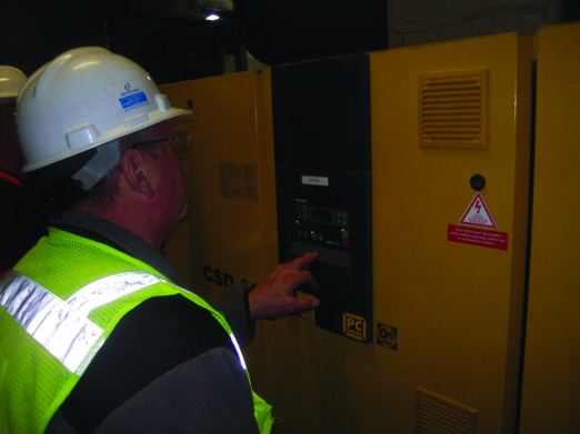 A worker in a white hard hat and yellow vest reviewing the control panel of an air compressor