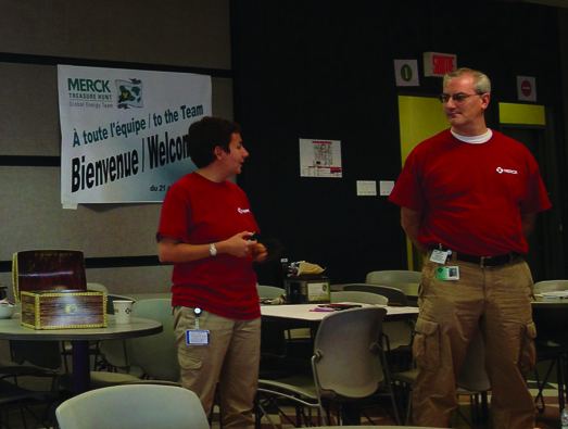 Two people in red shirts speaking at a kickoff meeting in front of a Merck welcome banner